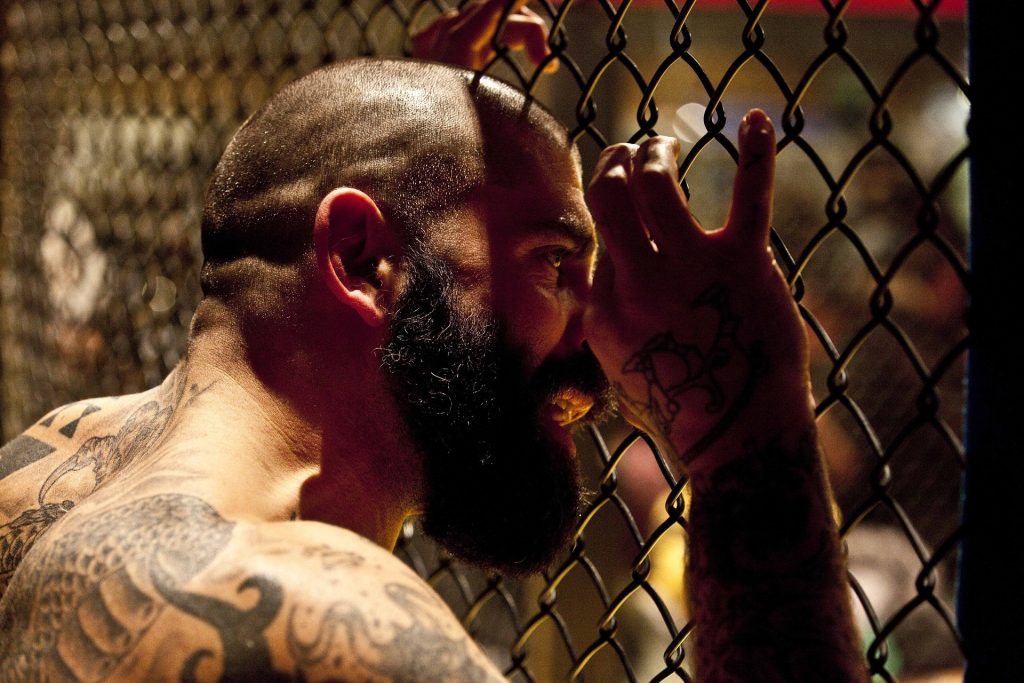 A man with a beard and tattoos leaning against a chain link fence