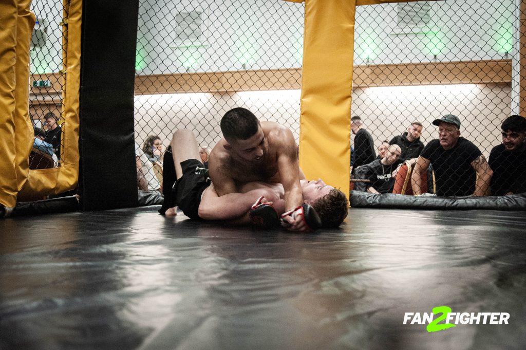 Two male fighters grappling on the mat in a cage at a mixed martial arts event, with spectators watching in the background.