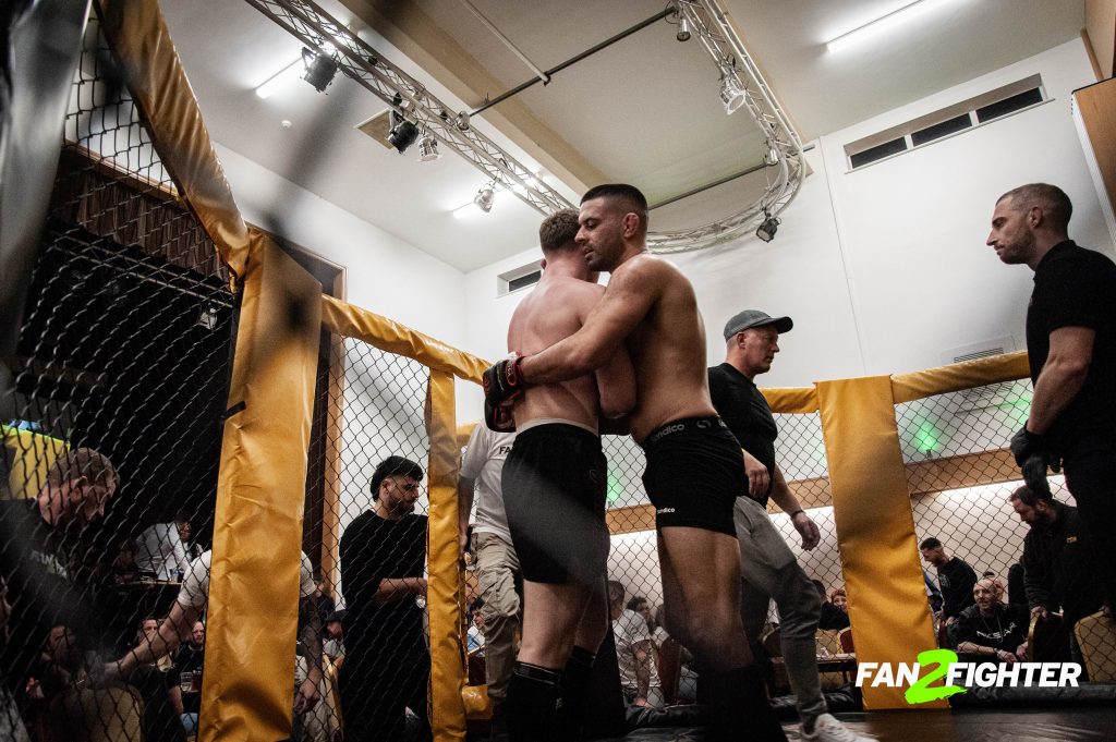 Two mixed martial arts fighters embrace in the ring, surrounded by a small crowd and a referee, in a gym with a chain-link fence.