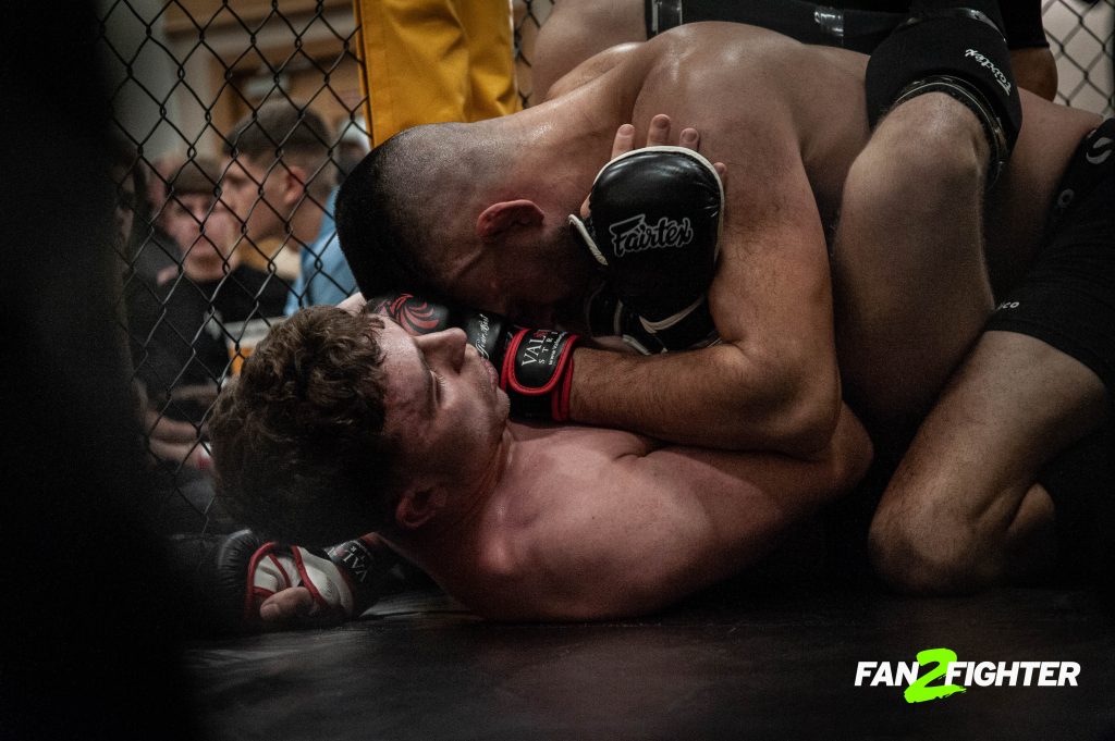 Two male mma fighters grappling on the ground in a caged ring, with one applying a chokehold to the other.