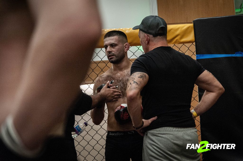 A shirtless male athlete with tattoos receives instructions from a coach in a gym, surrounded by other people in the background.