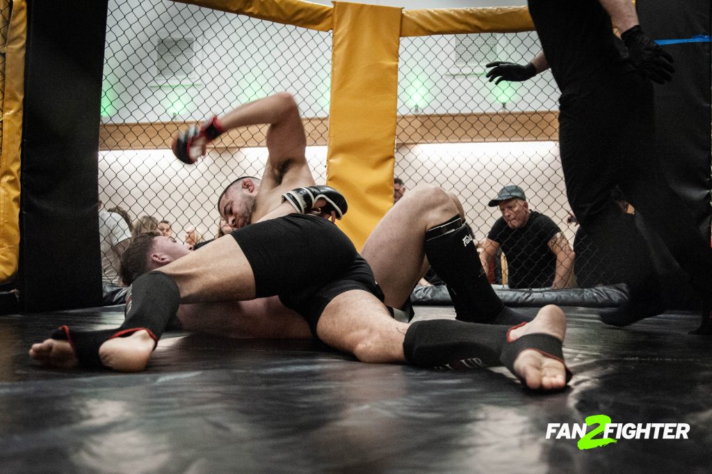 Two mma fighters grappling on the mat in a cage, with one in a dominant top position, as a referee watches closely.