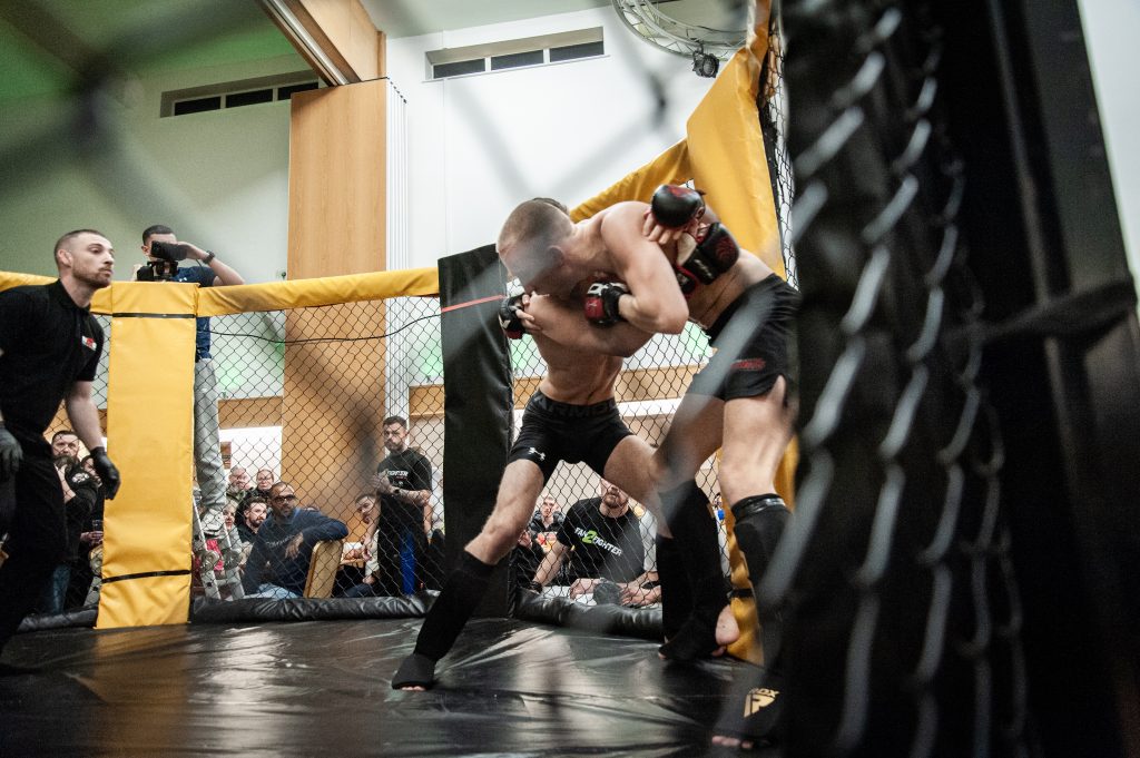 Two male mma fighters in a cage, one grappling the other against the fence, with a referee and audience in the background.