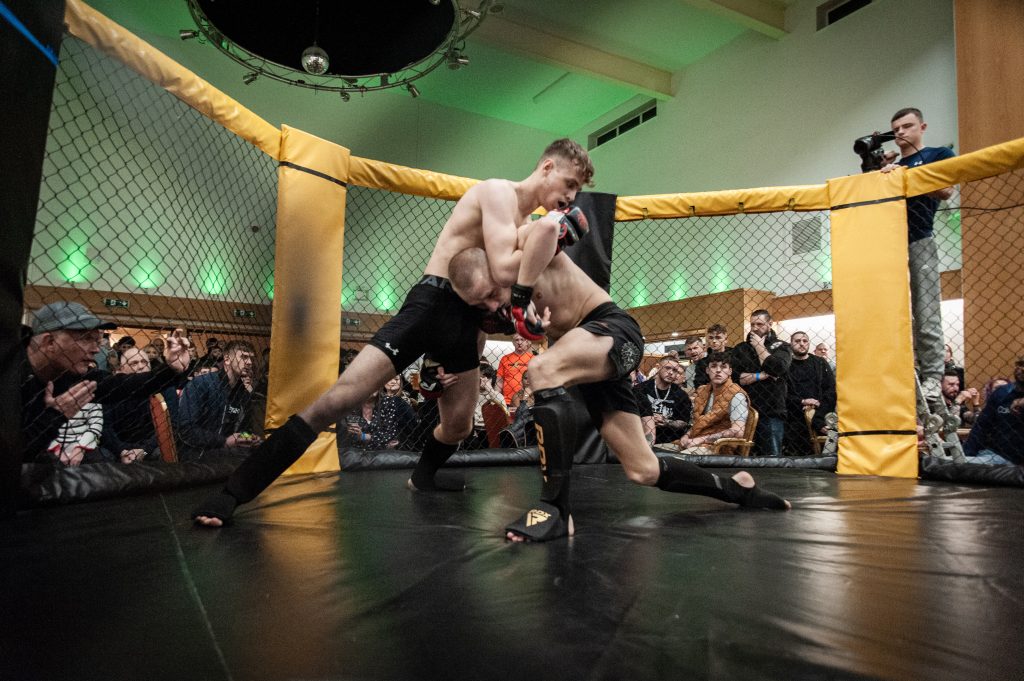 Two male mixed martial arts fighters compete in a cage, one landing a knee strike on the other, with an audience and a photographer observing.