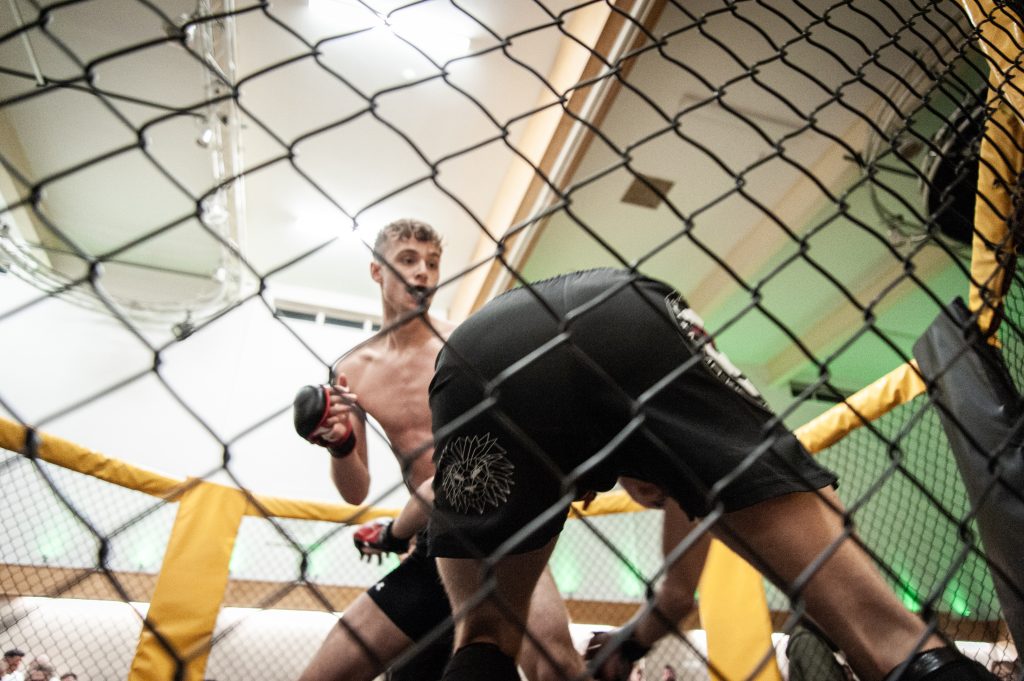 Two male mma fighters in a cage, one launching a knee strike towards the other who is bent forward, with a focus on the intensity of the match.