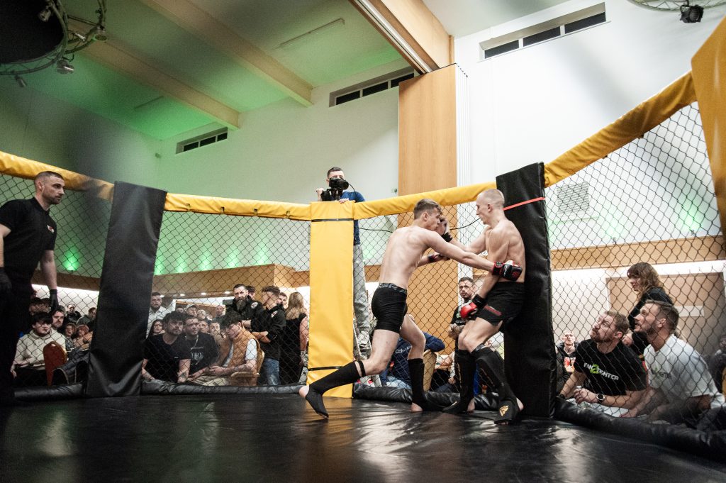 Two mixed martial arts fighters compete in a cage, surrounded by an attentive crowd, with a referee closely monitoring the action.