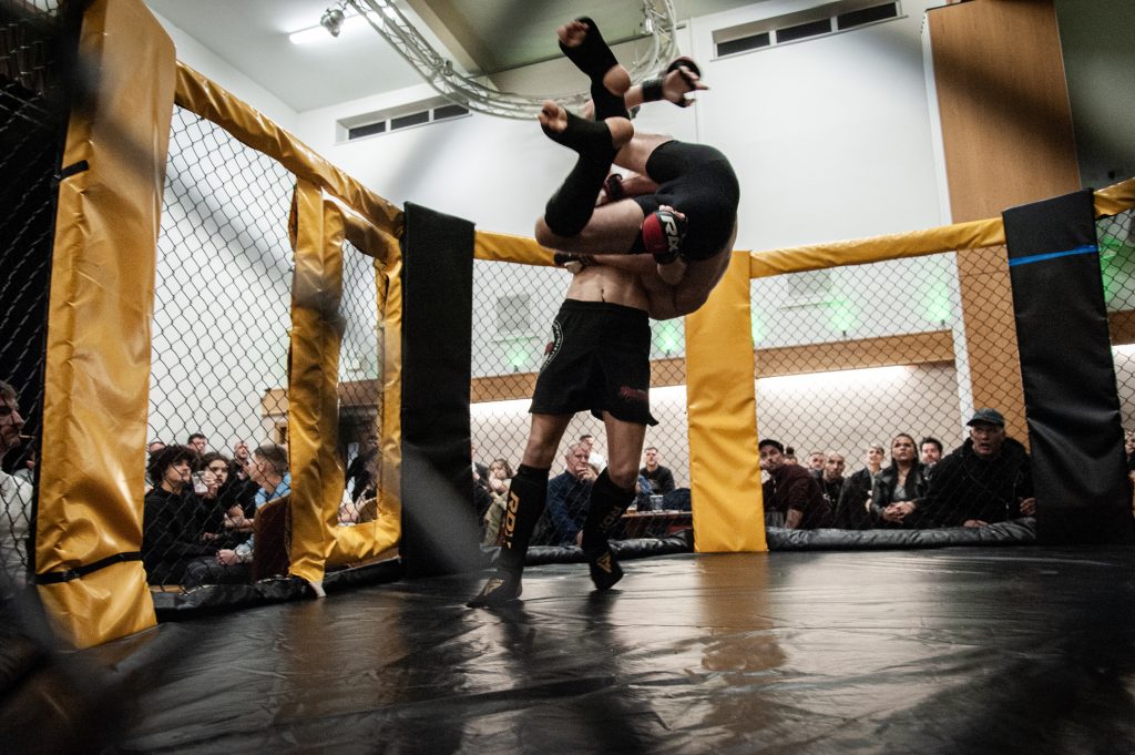 Two mixed martial arts fighters in a cage, one lifting the other upside down in a powerful move, with an attentive audience in the background.