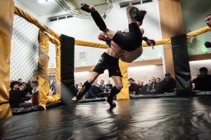 A mixed martial arts fighter executes an acrobatic flip inside a cage, as spectators watch closely from beyond the barriers.