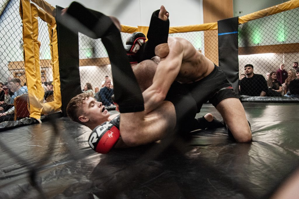 Two male mixed martial arts fighters grappling on a mat inside a cage, surrounded by spectators. one fighter is attempting a submission hold.