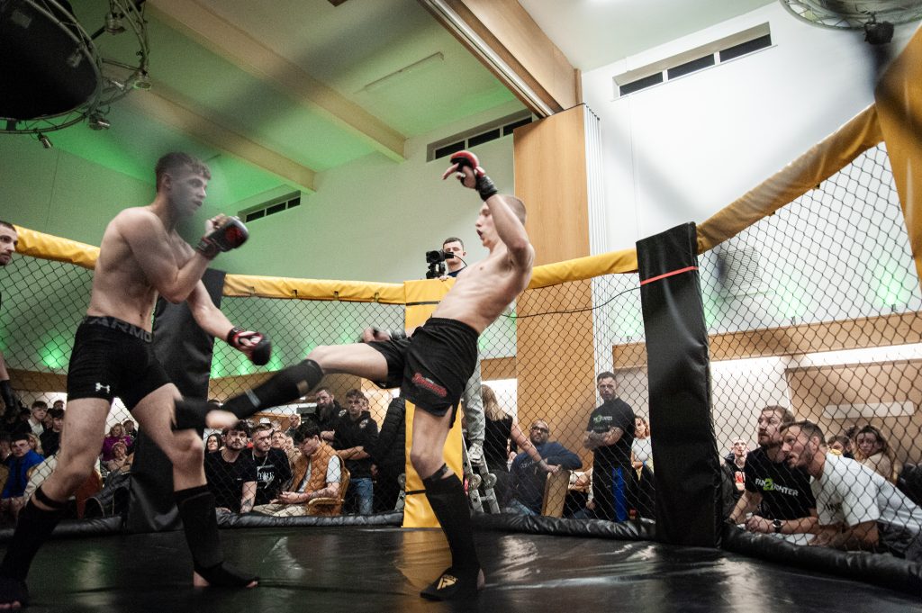 Two male mixed martial artists compete in a cage, with one fighter executing a high kick against his opponent. a focused crowd watches from behind the safety nets.