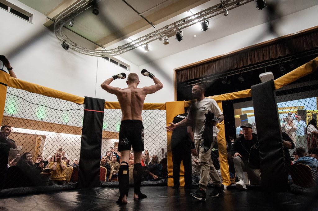 Two fighters prepare in an enclosed octagonal cage in a gym, surrounded by spectators. one fighter stretches while another chats with a coach, under bright lights and by projection screens.