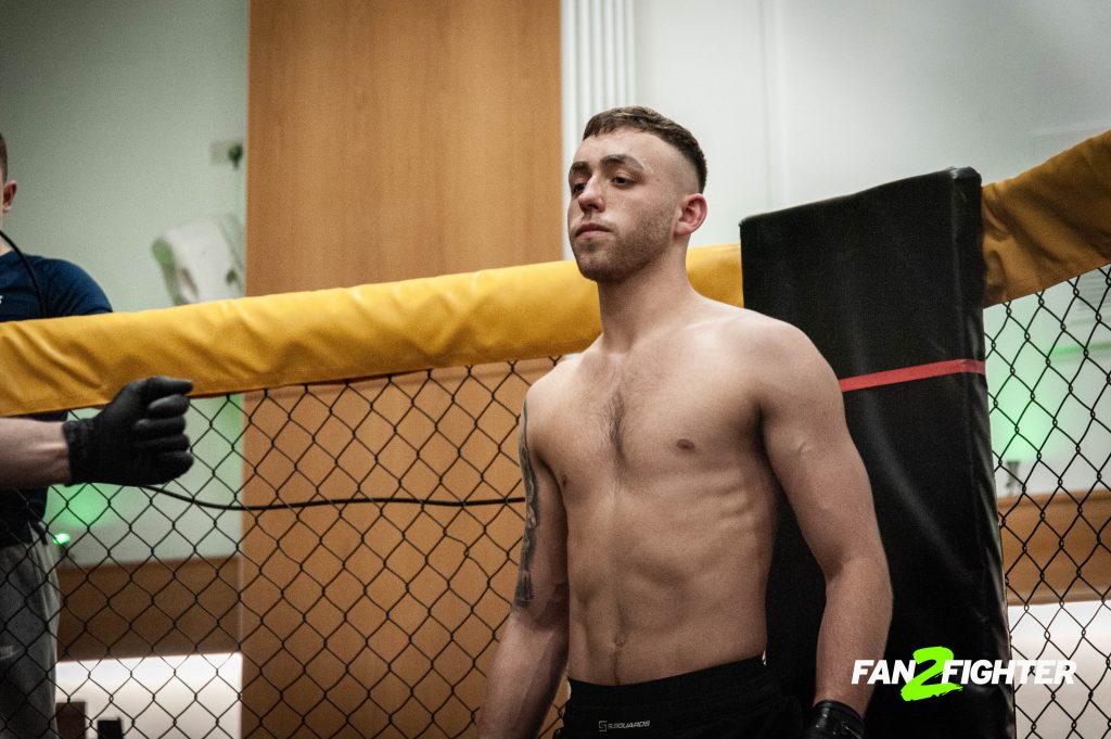 A muscular shirtless fighter stands before a wrestling cage, focused and ready for the match in a gym setting.