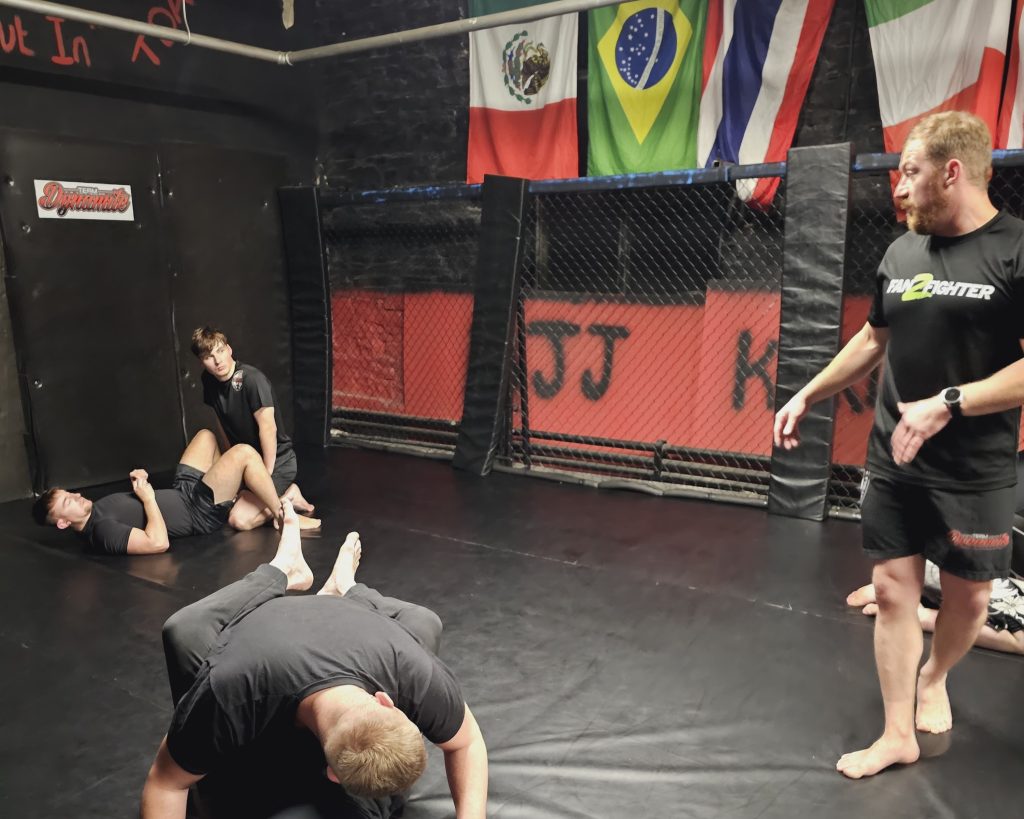 Martial arts gym with participants grappling, an instructor giving instructions, and flags in the background. Dim lighting.