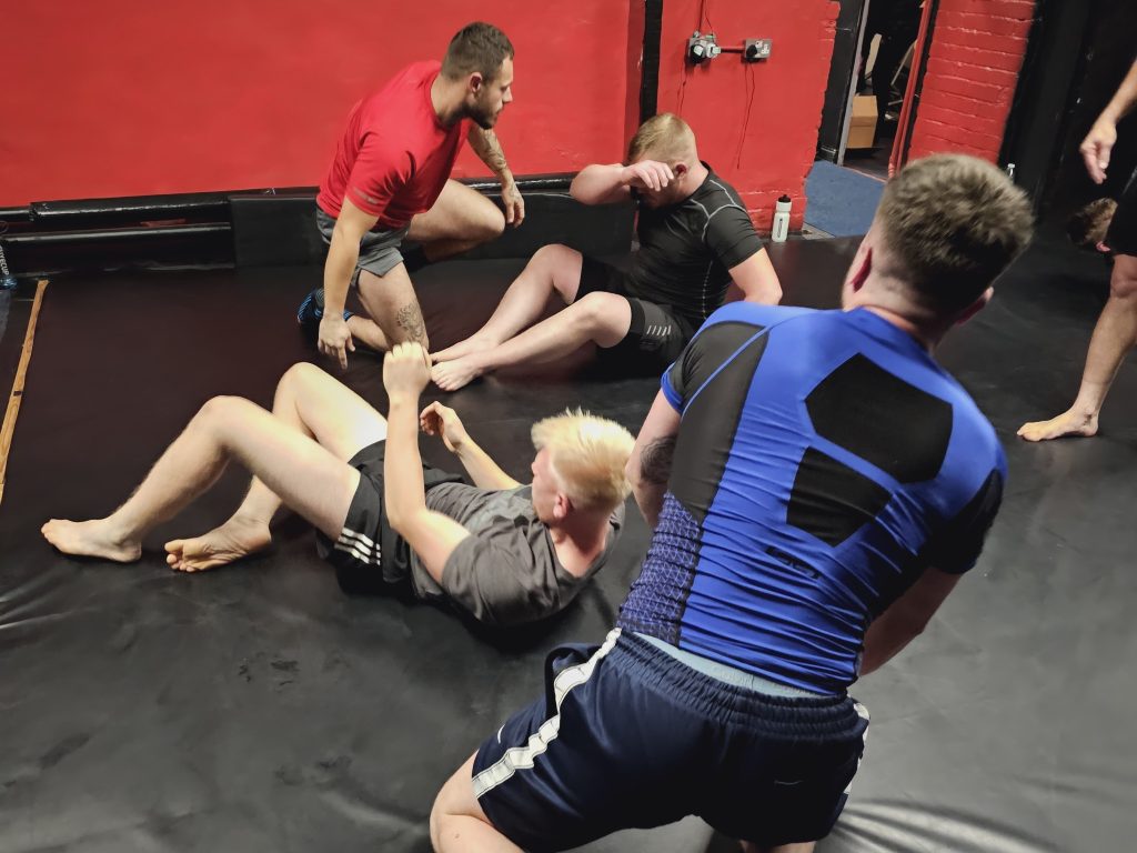 Five people grapple on a black mat in a gym with red walls and visible equipment. Two observe the action.