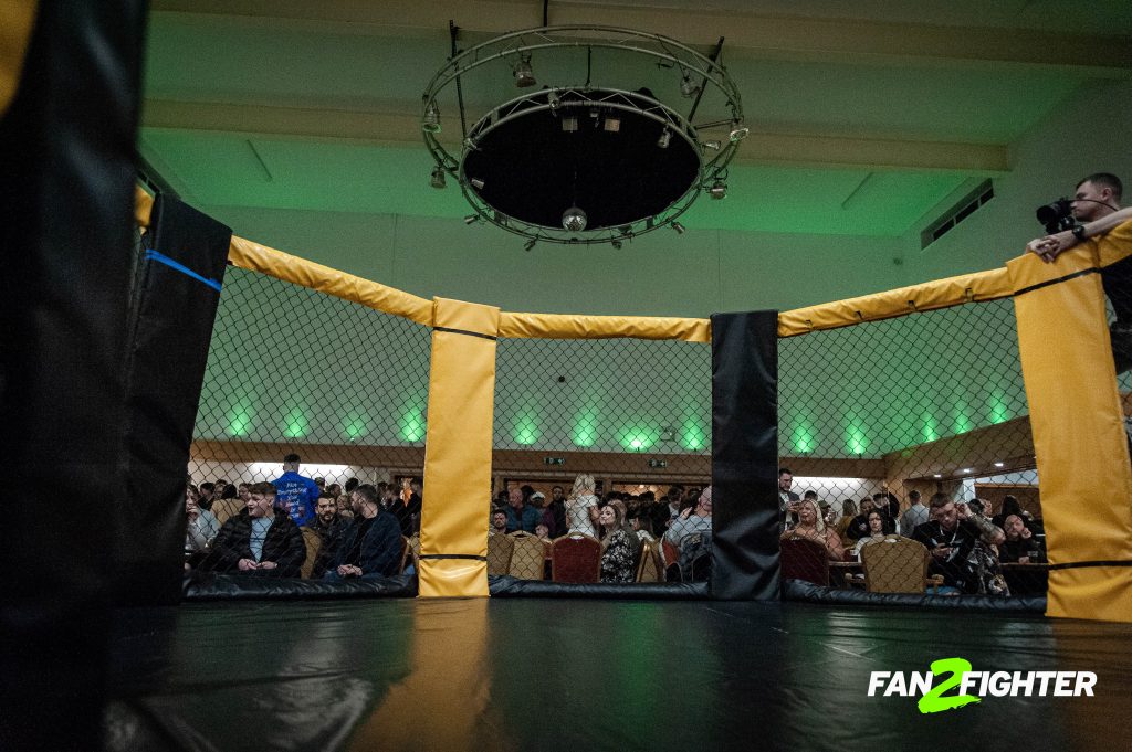 Low-angle view of a boxing ring with black and yellow walls, black mat, green-lit audience, and overhead lighting.