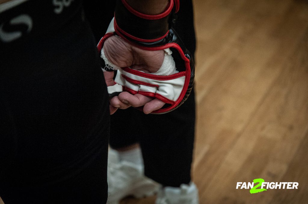 Close-up of a fighter's gloved hands, one black with red accents, gripping a white-gloved hand in a focused setting.