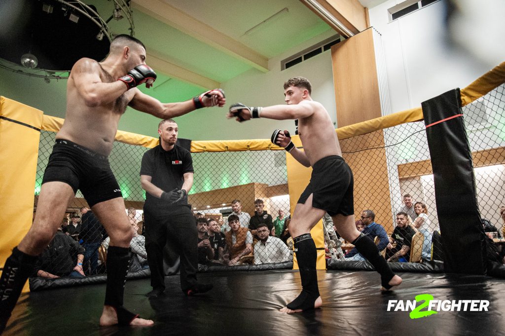 A mixed martial arts sparring match inside a cage, with fighters in black shorts and a referee overseeing the action.