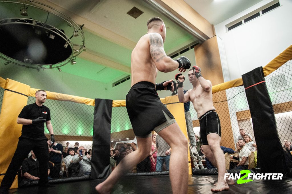 Two male fighters in combat stances inside an MMA ring, with a blurred audience and vibrant lighting.