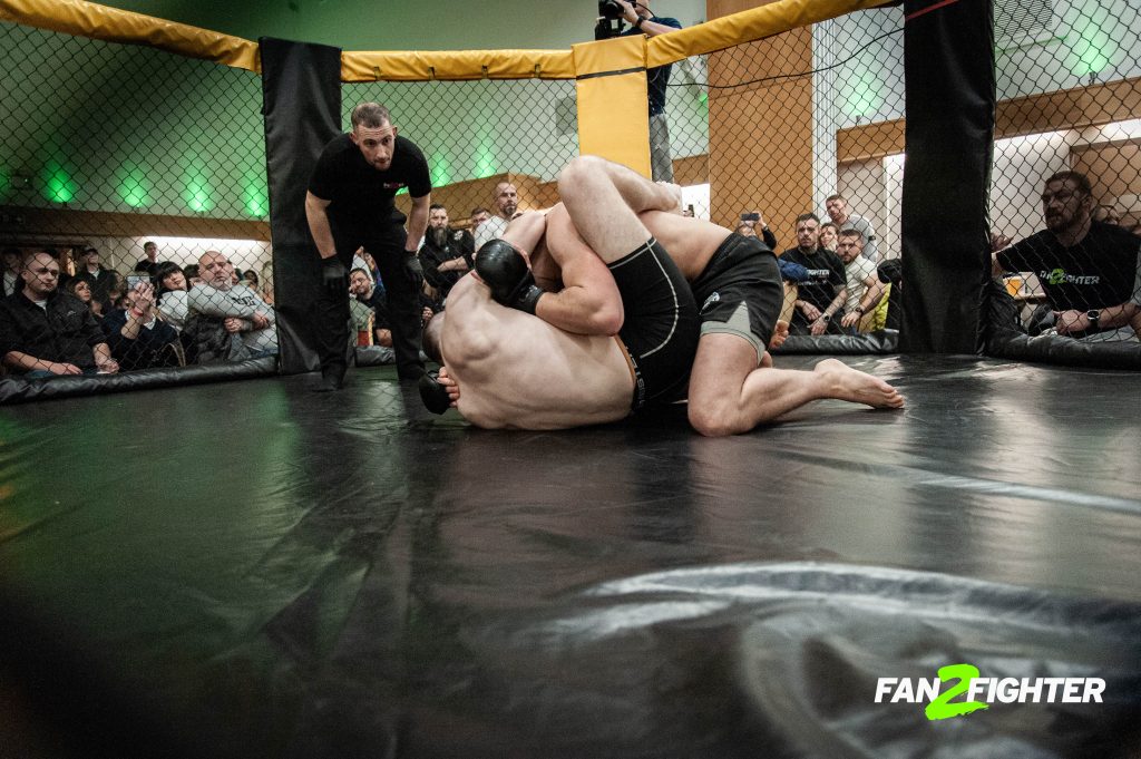 Two MMA fighters grapple on a black mat in the cage, with one in a dominant position and a referee nearby.