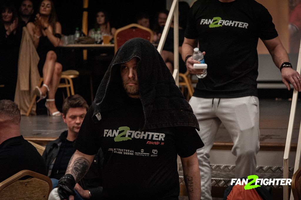 A man in a black T-shirt and towel walks towards the ring at a fighting event, with spectators in the background.