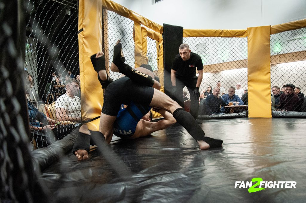 Mixed martial arts fighters in a cage, one on their back in blue, the other in black preparing a maneuver.