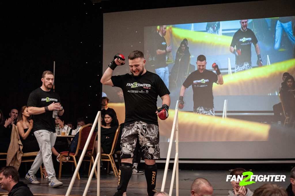 Male fighter in black shirt and patterned shorts raises hands in victory, with supportive companion and clapping audience.